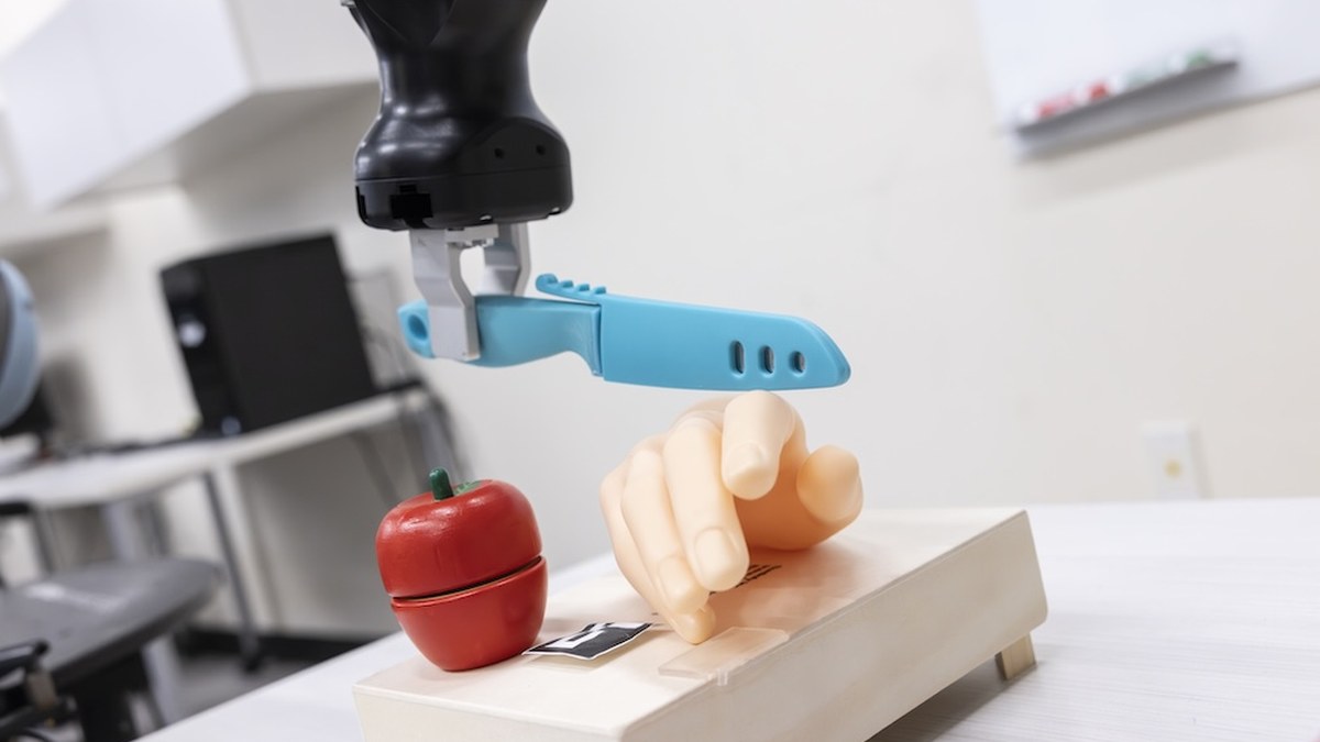 Un bras robotisé tient un couteau au-dessus d'une pomme en plastique et d'une main factice